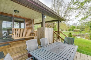 A covered outdoor deck with wooden furniture, a hanging swing, and a view of a pond and trees. - Lakefront Getaway w/ Fire Pit in Akron (Akron)
