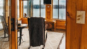 Interieur van een houten hutje met een groot raam dat uitkijkt over een meer, een houten bureau en een stoel.