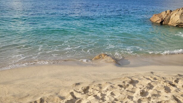 A sandy beach with clear blue water and a rocky outcrop.