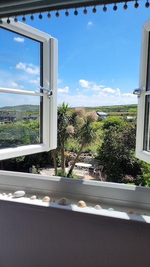 An open window with a view of a green landscape, a stone wall, and a white chair outside.