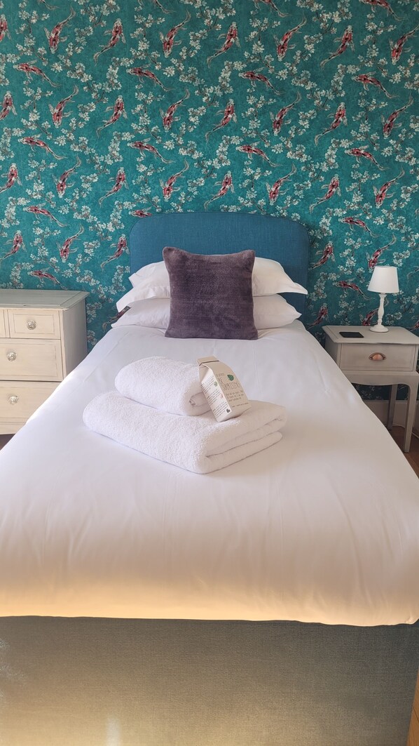 A neatly made bed with white linens, a purple pillow, and a folded towel on a bed in a room with teal floral wallpaper.