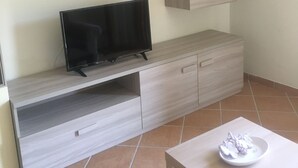 Ein modernes Wohnzimmer mit einem Flachbildfernseher auf einem Holz-TV-Möbel, einem kleinen Schrank mit Spiegel und einem Fliesenboden.
