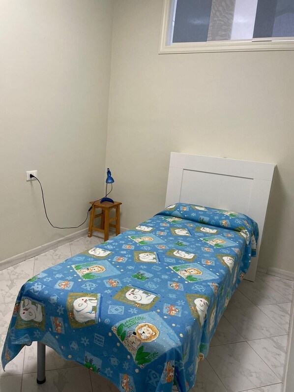 A small, neatly arranged bedroom with a bed covered in a blue bedspread featuring cartoon characters, a white headboard, and a small wooden stool with a lamp.