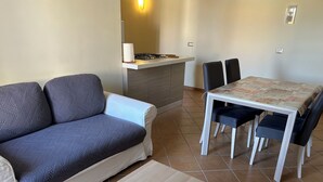 Ein Wohnzimmer mit einer Couch, einem Esstisch mit Stühlen, einem kleinen Holztisch und einer Küchenzeile mit Gasherd und Arbeitsplatte.