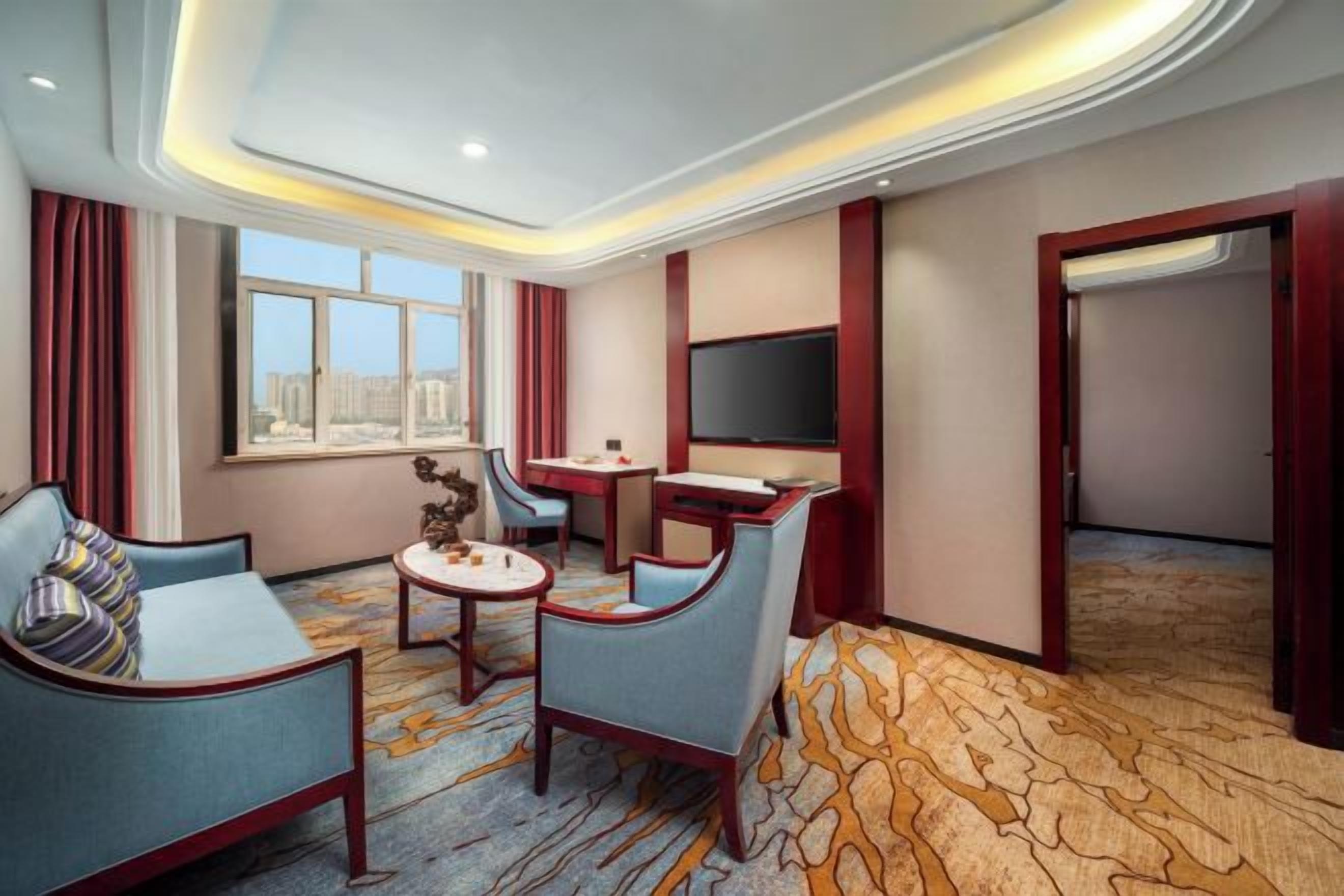 A hotel room with a sofa, armchair, small table, TV, and a view of the city.