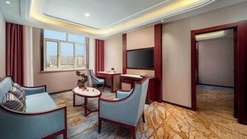 A hotel room with a sofa, armchair, small table, TV, and a view of the city.