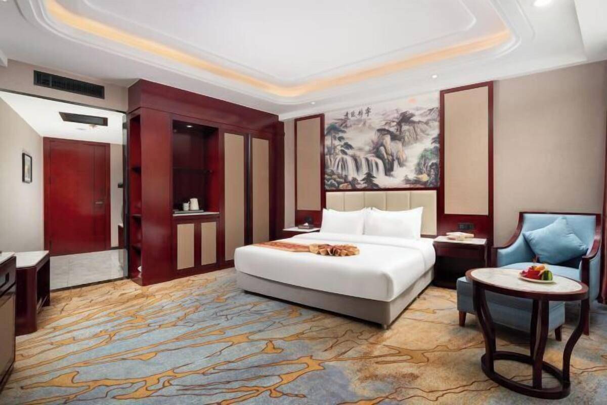 A hotel room with a bed, a desk, a chair, a small table, and a painting on the wall.