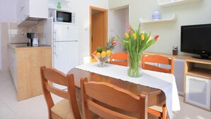 A compact kitchen with a microwave, refrigerator, and coffee maker. There is a dining table with chairs and a vase of flowers. A television is on a stand to the right. - Apartments Roščić (Podgora)