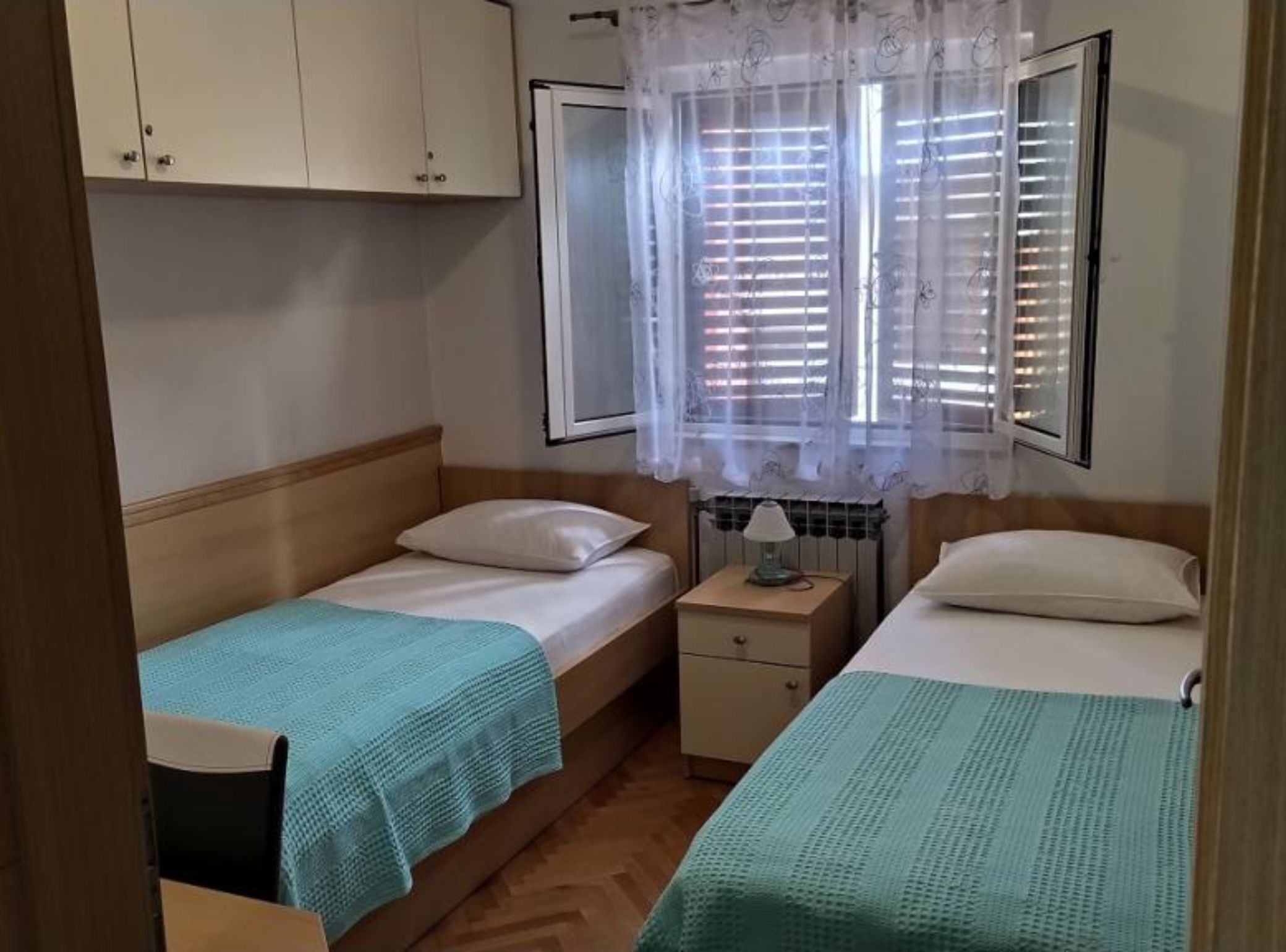 A small, tidy bedroom with two single beds, a nightstand with a lamp, and a window with blinds.