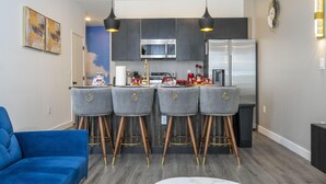 Une cuisine moderne avec des meubles de couleur sombre, un four à micro-ondes en acier inoxydable et un réfrigérateur. On y trouve quatre tabourets de bar avec des détails dorés et un canapé bleu dans l’espace salon.