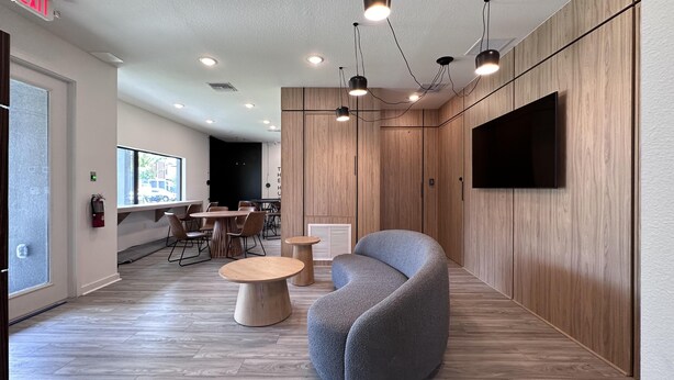 Un espace de bureau moderne avec un canapé gris recouvert de tissu, des tables rondes en bois et un grand téléviseur à écran plat fixé au mur.