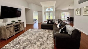 A spacious living room with a large flat-screen TV, a dark leather sofa, and a patterned rug. - Heated pool, pool table, heart of Wellington! (Wellington)