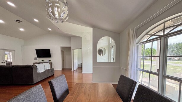 A spacious living room with a large wooden dining table, grey upholstered chairs, a grey sofa, a flat-screen TV, and a large arched window offering a view of the outdoors. - Heated pool, pool table, heart of Wellington! (Wellington)