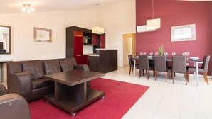 Un salón moderno con un sofá de cuero marrón, una mesa de centro oscura y una alfombra roja. Hay un comedor con una mesa preparada para cuatro personas y sillas. La habitación tiene una pared de acento roja y una lámpara en el techo.