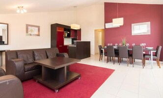 Un salón moderno con un sofá de cuero marrón, una mesa de centro oscura y una alfombra roja. Hay un comedor con una mesa preparada para cuatro personas y sillas. La habitación tiene una pared de acento roja y una lámpara en el techo.