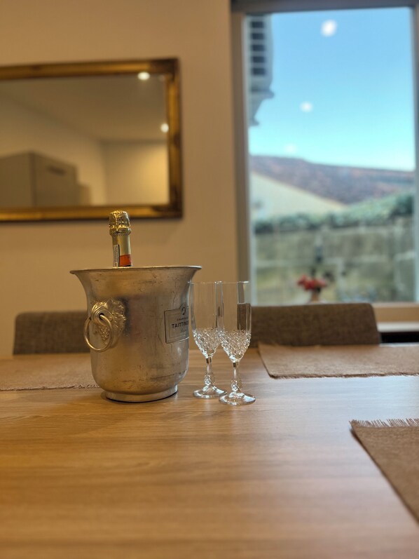 A metallic champagne bucket with a bottle on a wooden table, two empty wine glasses, a framed mirror, and a window with a view of a landscape.