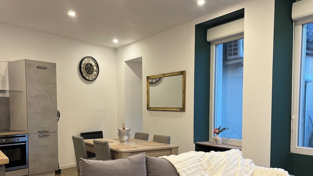 A modern living room with a grey sectional sofa, a dining table with chairs, a kitchen area with a built-in oven, a wall-mounted clock, and a window with a view.