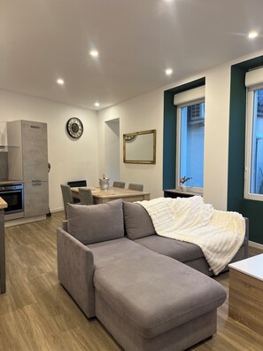 A modern living room with a grey sectional sofa, a dining table with chairs, a kitchen area with a built-in oven, a wall-mounted clock, and a window with a view.