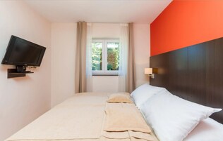 Habitación de hotel con cama, televisor montado en la pared, ventana con cortinas y vista a zonas verdes.