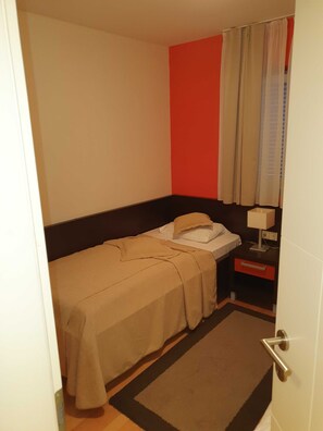 A small, single-bed room with a bed, nightstand, lamp, and a door. - Marina Portorož Family Friendly Apartment 5 (Piran)