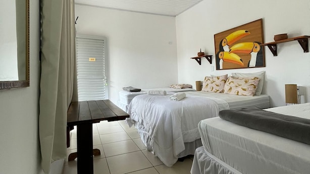 A hotel room with two beds, a wooden bench, a painting of toucans, and a window with blinds. - Chill Inn Pousada Paraty (Paraty)