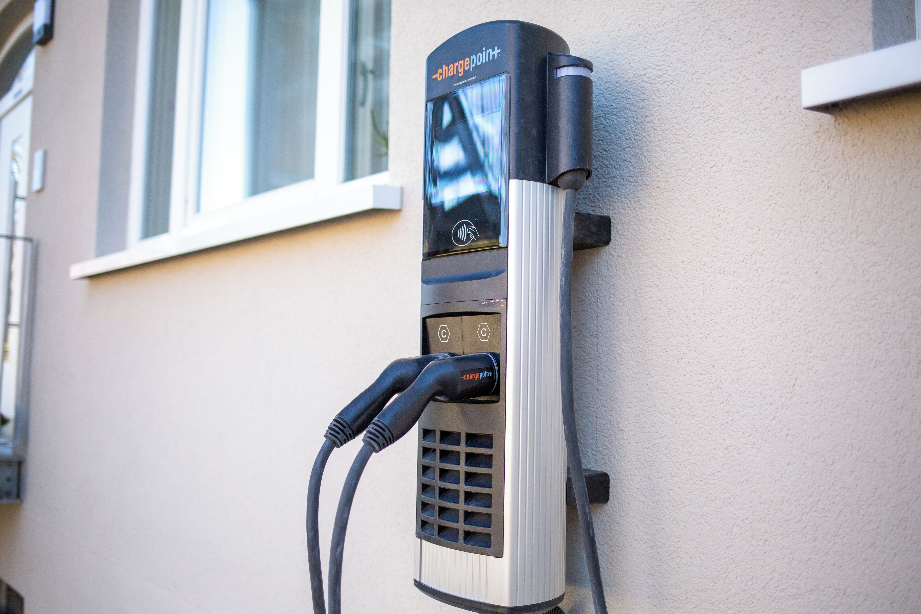 Stazione di ricarica per veicoli elettrici montata a parete, con display digitale, due prese di ricarica e un cavo collegato a un veicolo.