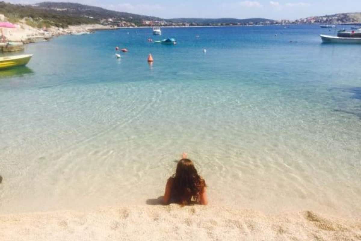 A beach with clear water, boats, and a person sitting on the sand.