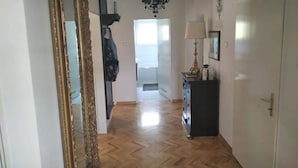 A hallway with a wooden floor, a large ornate mirror, a chandelier, and a door leading to another room.