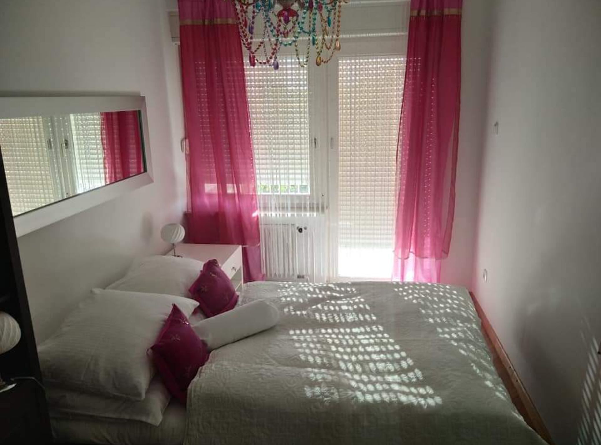 Un dormitorio con cama, almohadas, una mesita de noche y una ventana con cortinas.