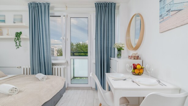 Ein Hotelzimmer mit Bett, Esstisch mit Stühlen, einer kleinen Küche und einem Balkon mit Vorhängen.