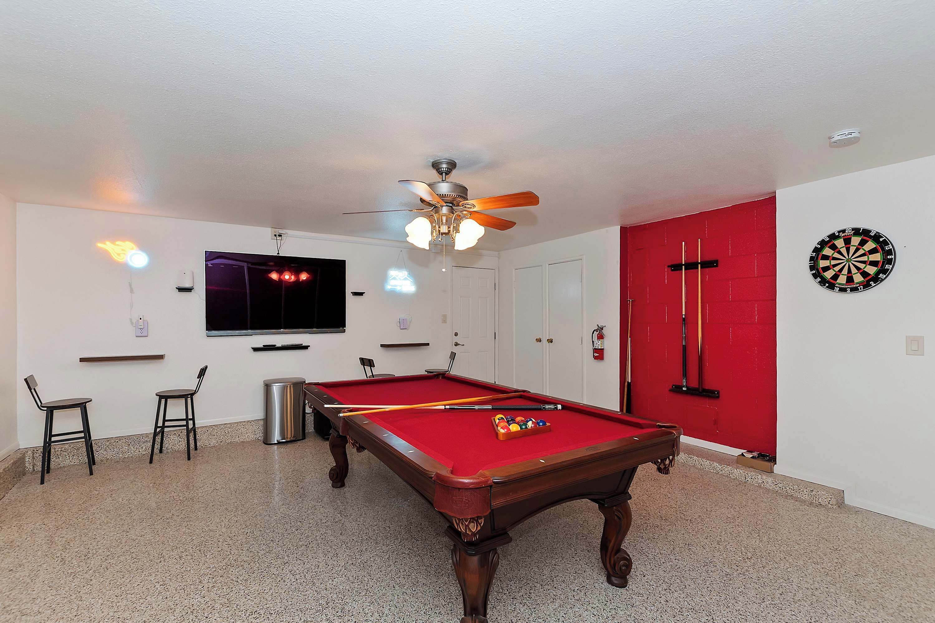 A pool table with a red felt surface, a ceiling fan, and a dartboard on the wall.