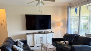 Un salon avec deux canapés noirs, une table basse blanche, un ventilateur de plafond, un téléviseur à écran plat et une fenêtre avec des stores.