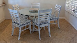 Un coin repas avec une table ronde en verre, entourée de quatre chaises blanches dotées de coussins à motifs. Un lustre est suspendu au plafond et les murs sont peints en blanc.