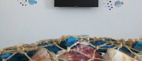 A television mounted on a wall with a netted basket of seashells below it.