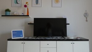 Une pièce avec un téléviseur à écran plat posé sur un meuble, une armoire blanche avec des tiroirs, une petite étagère avec une plante et des fleurs, et trois tableaux encadrés accrochés au mur.