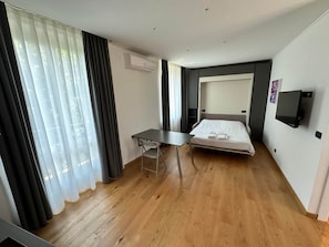 A hotel room with a bed, a desk, a chair, an air conditioning unit, a TV, and a window with curtains.