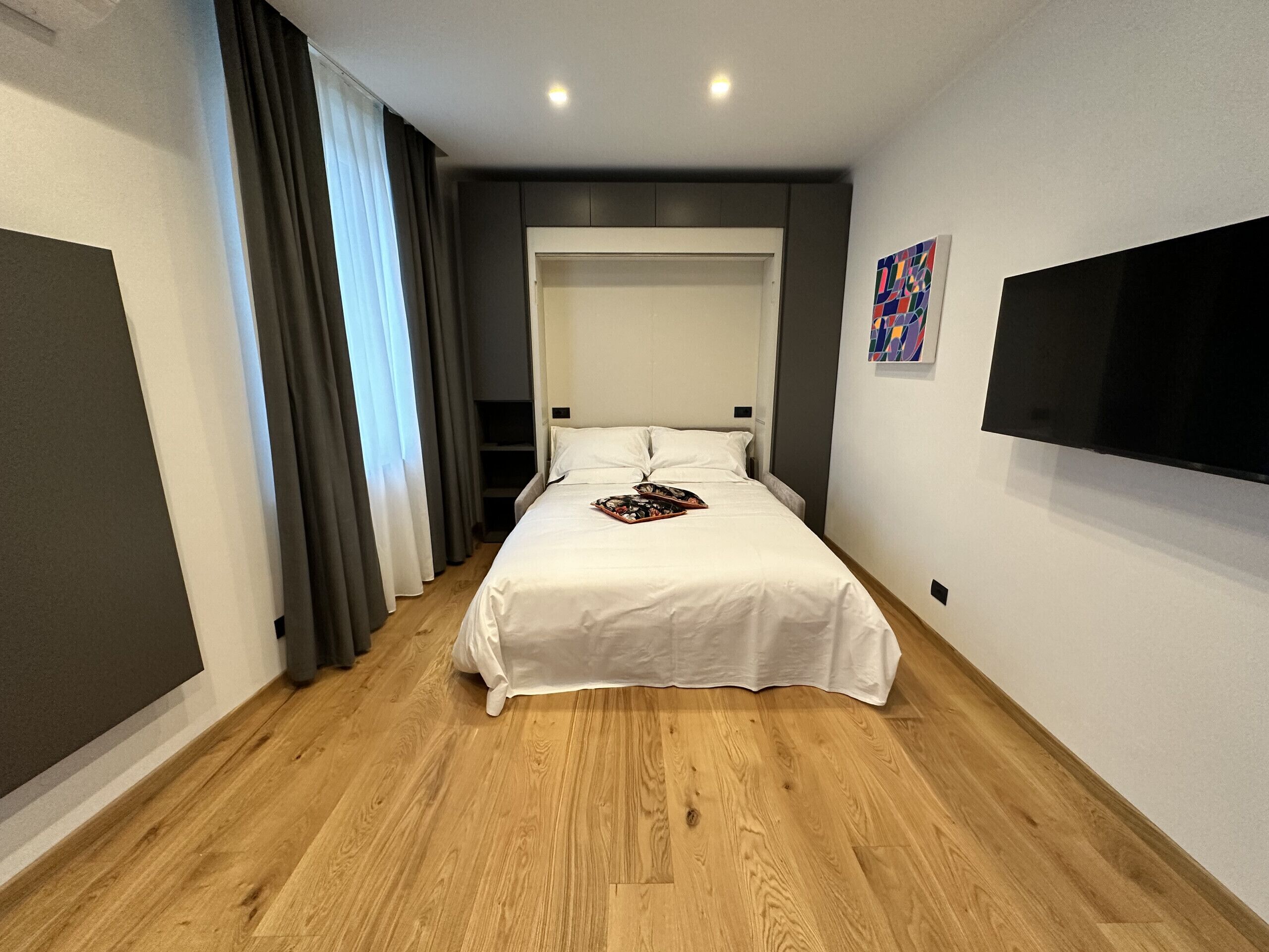 A modern hotel room with a large bed, a flat-screen TV mounted on the wall, and a wooden floor.