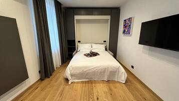 A modern hotel room with a large bed, a flat-screen TV mounted on the wall, and a wooden floor.
