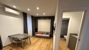 Ein modernes Wohnzimmer mit Holzboden, einem grauen Sofa mit orangefarbenem Kissen, einem Esstisch mit Stühlen und einer eingebauten Klimaanlage.