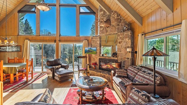 Un vaste salon doté d’une cheminée en pierre, d’un plafond en bois et de grandes fenêtres offrant une vue sur l’extérieur.