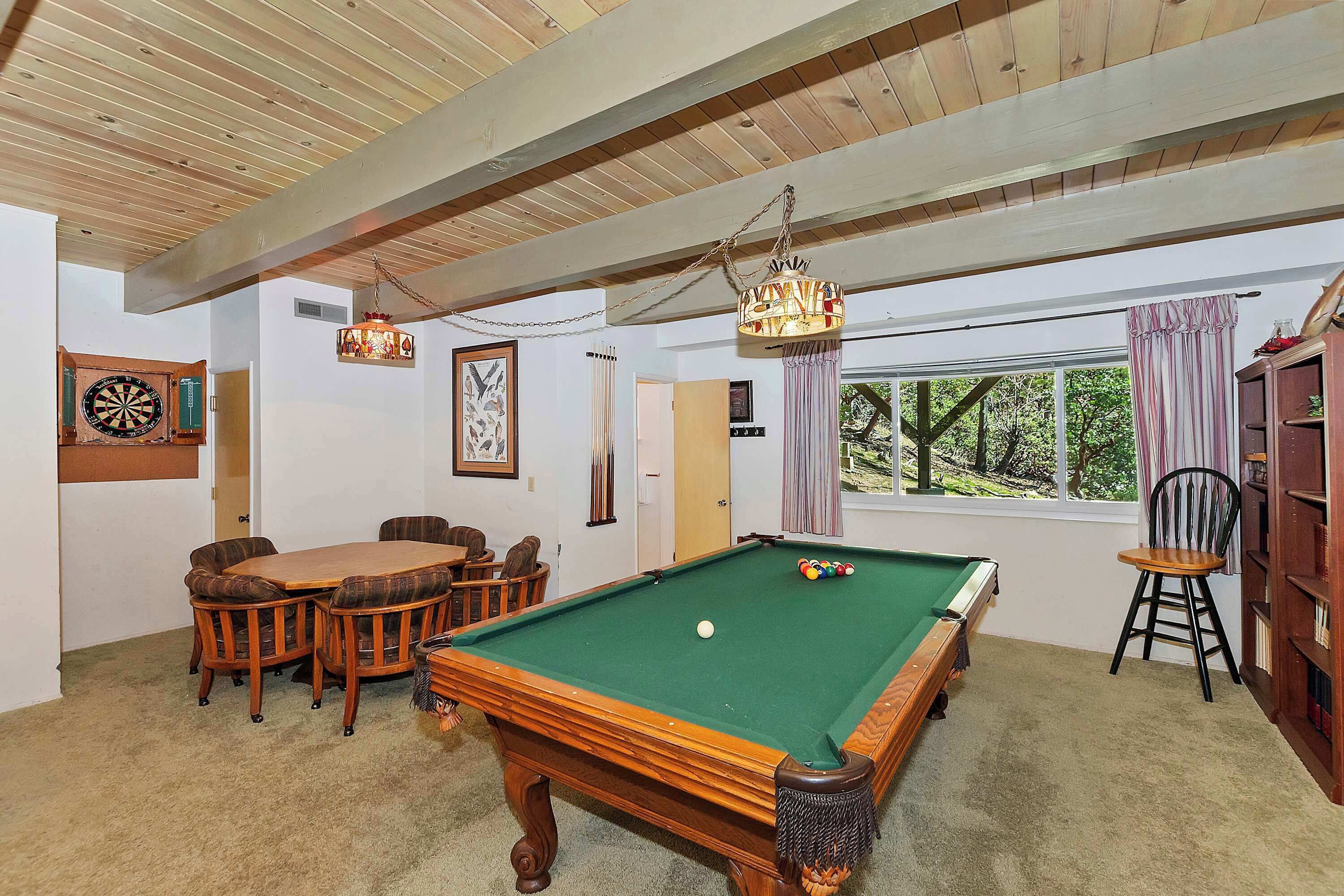 A room with a pool table, a dartboard, a dining table with chairs, and a bookshelf.