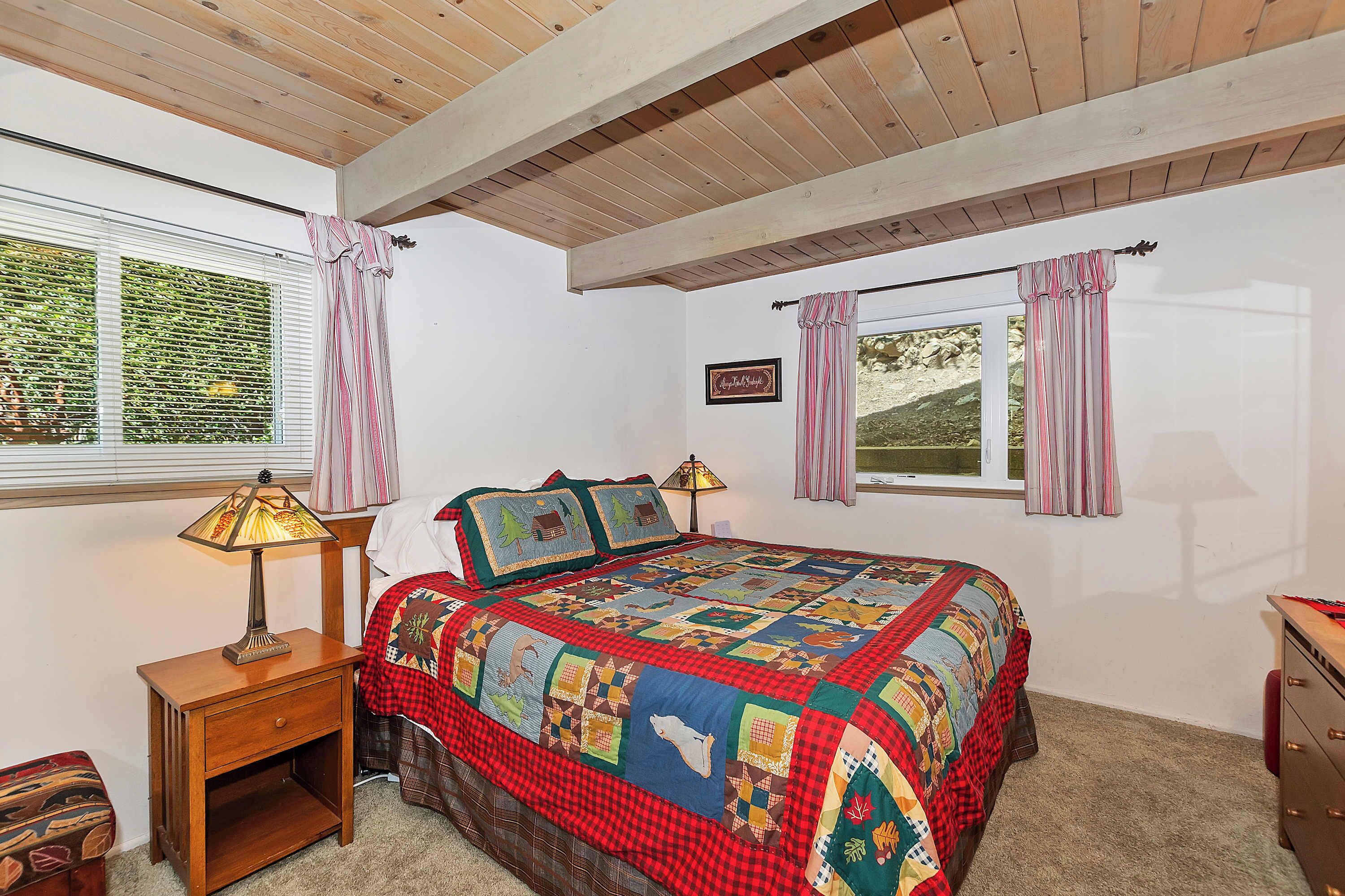 Une chambre avec un plafond en bois, un grand lit recouvert d’une couette aux couleurs vives, deux lampes de chevet et une table de chevet avec une lampe.
