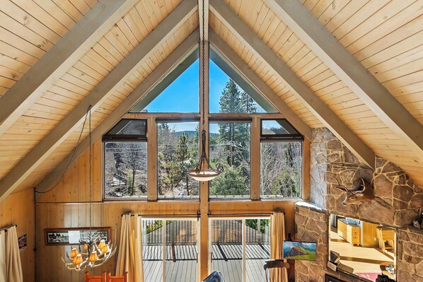 A high-ceilinged room with wooden beams, large windows, a stone fireplace, and a chandelier.