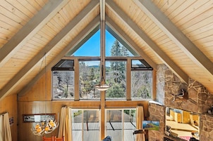 A high-ceilinged room with wooden beams, large windows, a stone fireplace, and a chandelier.
