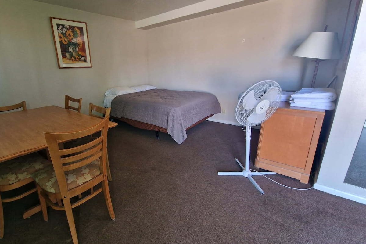 A small room with a wooden table and chairs, a bed with a gray blanket, a standing fan, and a lamp.