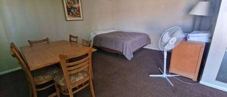 Ein kleiner Raum mit einem Holztisch und Stühlen, einem Bett mit grauer Decke, einem Standventilator und einer Lampe auf einem Schrank.