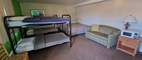 Una cama litera con una cama inferior, una cama individual, un sofá a rayas, una mesita de noche de madera con un microondas y una lámpara.