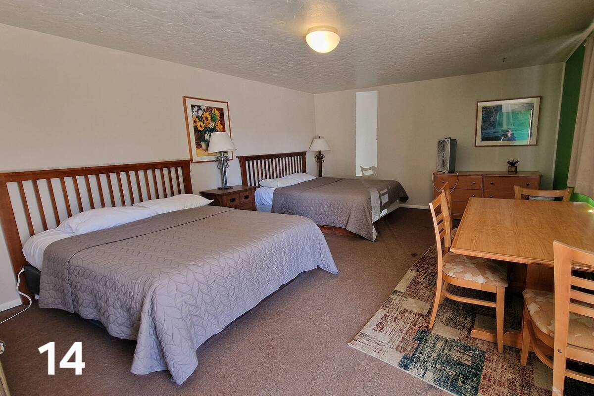 A hotel room with two single beds, a wooden dining table with chairs, a small desk, a fan, and a painting on the wall.