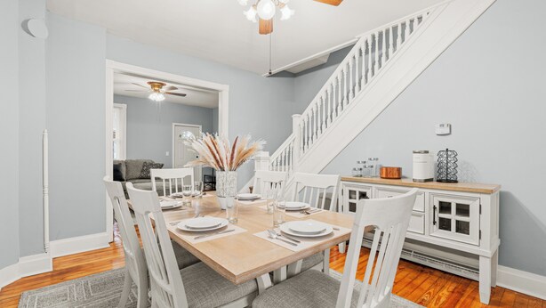 A dining area with a wooden table set for four, white chairs, a ceiling fan, and a staircase in the background.
