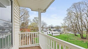 A covered porch with a wooden deck and white railings, overlooking a grassy area with houses and trees.